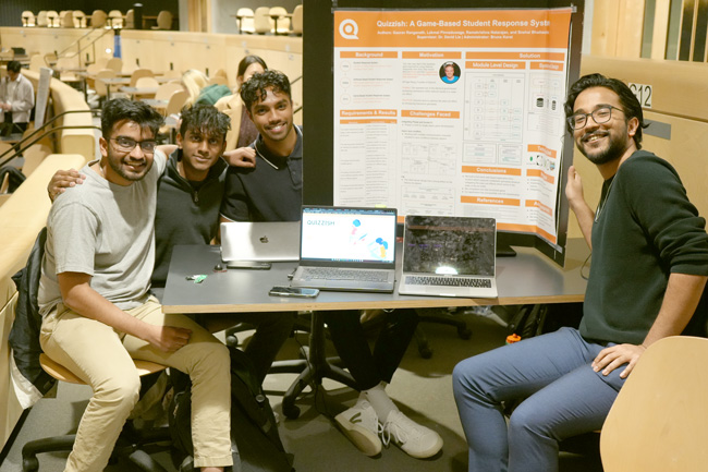 Four smiling students sitting at table with project board and laptops between them