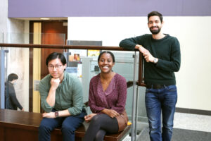 Two sitting students, one standing, in Bahen Centre after class, smiling at the camera