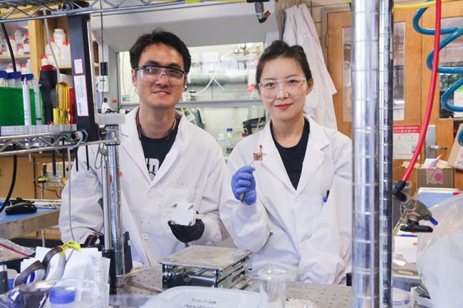 On the left Rui Kai (Ray) Miao stands in a lab holding a small square device. Beside him on the right is Dr. Mengyang Fan who is holding up a small chip-like device with tweezers.