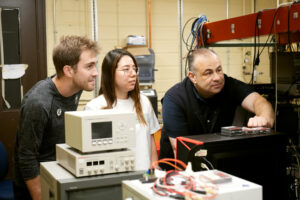 Two students behind equipment in lab, professor pointing to a monitor