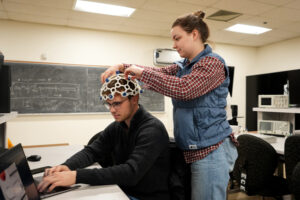 One student, standing, tightens electrodes on helmet worn by another, seated