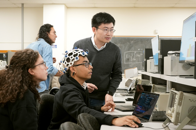 Students and professors looking at monitor while one student is connected by electrodes to a headset