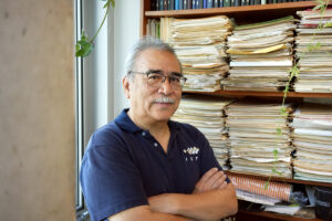 Professor with arms crossed, standing before shelf with stacks of paper