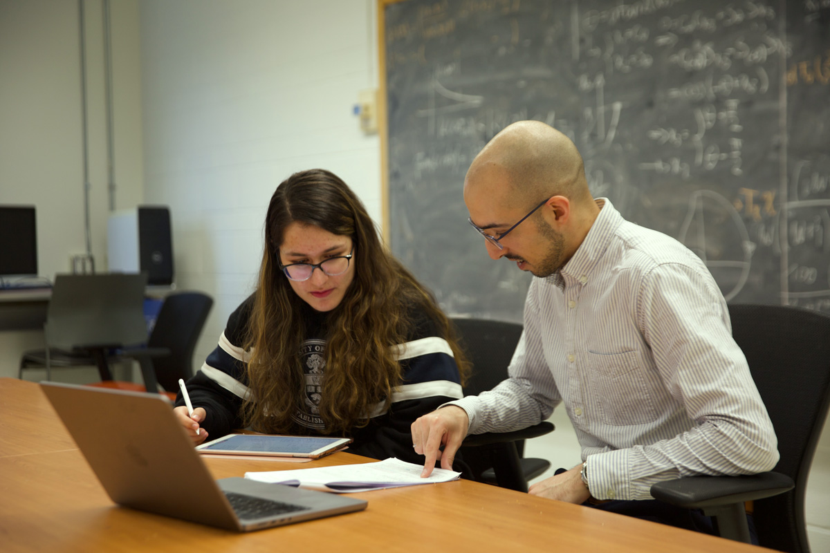 Seated student listens to professor, who is pointing to a paper, chalkboard in the background